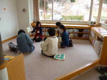 本棚に囲まれたブースでお母さんの膝に抱っこされた4組の赤ちゃんが絵本の読み聞かせを聞いている写真
