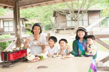 ご夫婦と3人のお子様の家族連れが野外のテーブルでバーベキューを楽しんでいる写真