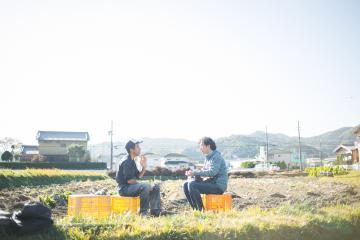 畑の中で黄色のコンテナを椅子にして座り、男性2人が向き合って話をしている写真