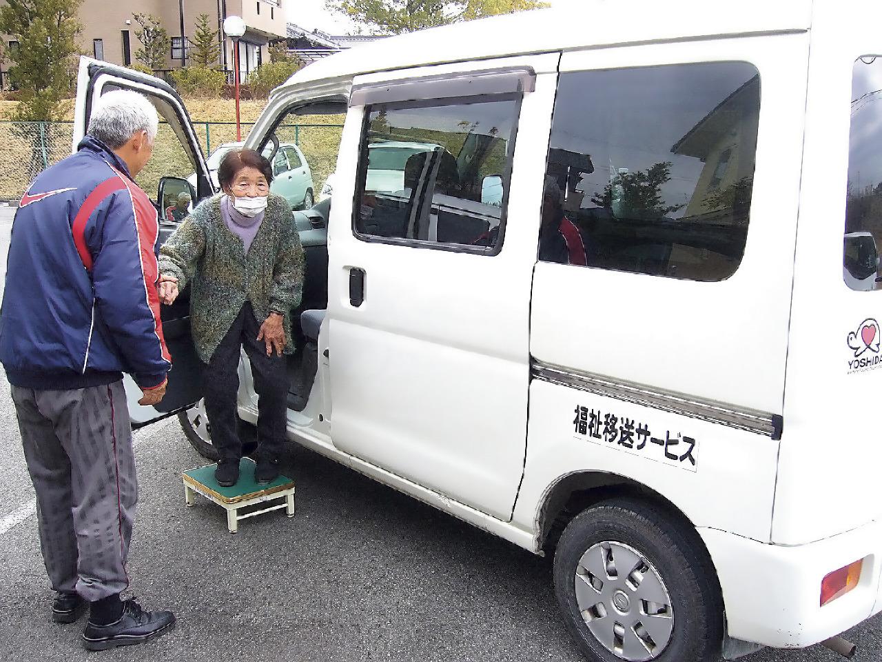 高齢者の方がスタッフの方のサポートを受けて白のワゴン車から降りている写真